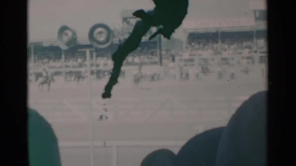 1960 Cheyenne rodeo footage showing a horse and rider mid-action beyond foreground spectator heads and arena fencing.