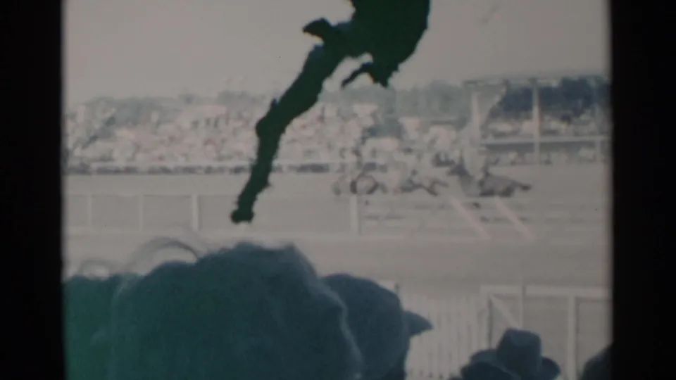 Cheyenne rodeo home-movie frame showing the arena, distant horse action, grandstands, and dark foreground spectator silhouettes.
