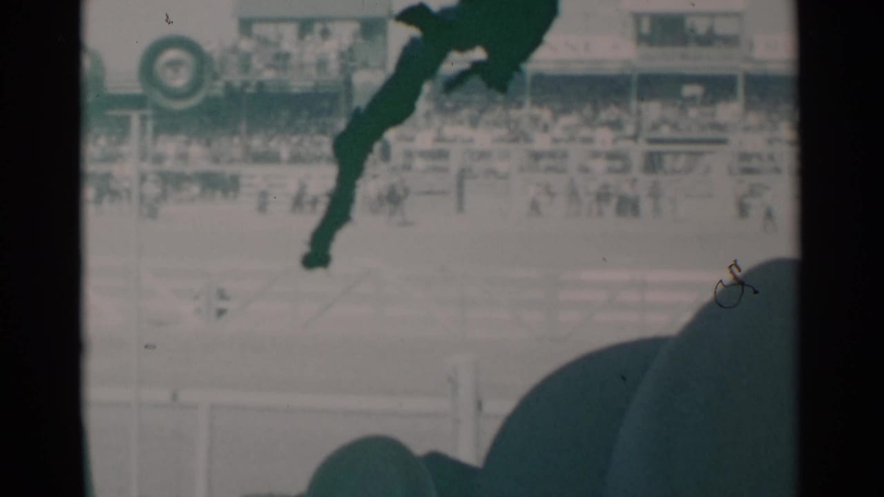 Rodeo arena with rider and bronco, grandstand crowd, signage, and a dark foreground head.