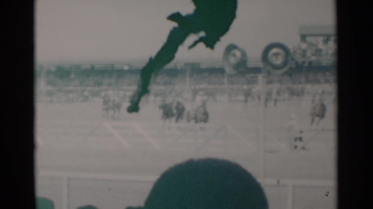 1960 Cheyenne, Wyoming home-movie frame showing horses and riders in a dusty arena, with bleachers and a dark spectator silhouette in the foreground.