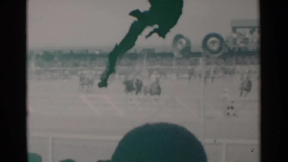 1960 Cheyenne, Wyoming home-movie frame showing horses and riders in a dusty arena, with bleachers and a dark spectator silhouette in the foreground.