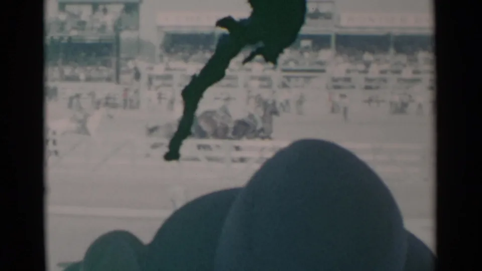 Home-movie view of a 1960 Cheyenne arena where multiple horses and riders appear near the track, with bleachers and a foreground spectator silhouette.