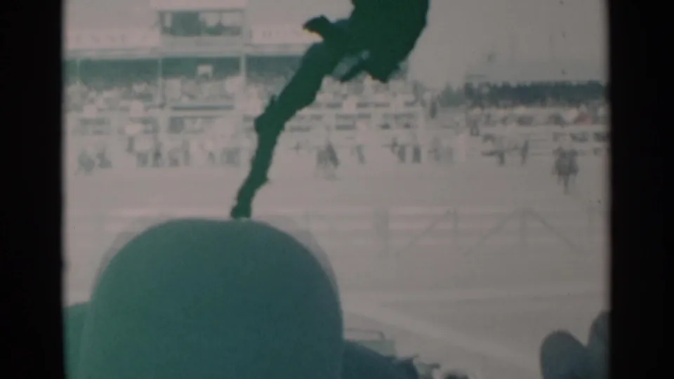The 1960 Cheyenne horse race remains visible beyond a foreground spectator hat, with railings, riders, and bleachers softened by 8mm film texture.