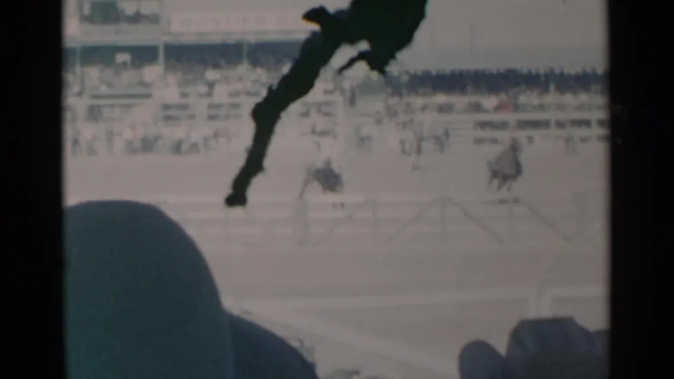 1960 Cheyenne rodeo footage shows riders separated across the arena, with spectator heads at the bottom edge and stands behind them.