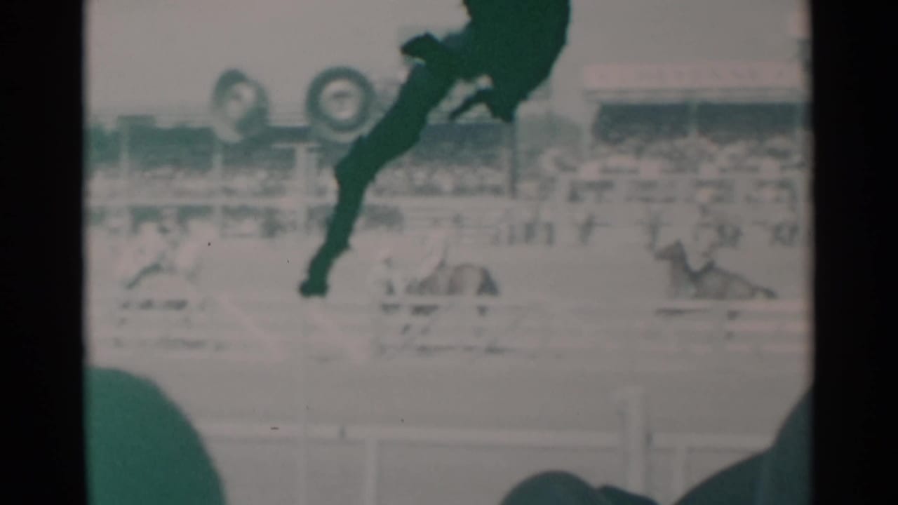 Horse and rider pass a dirt track while spectators sit in the foreground.