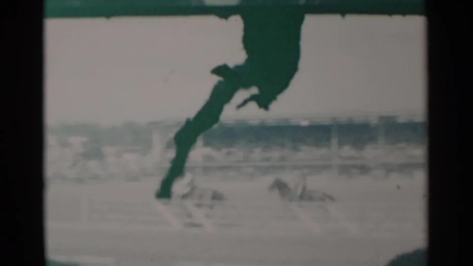 A dark horse-and-rider silhouette moves through the upper frame, with the pale dirt track and distant stands behind it.