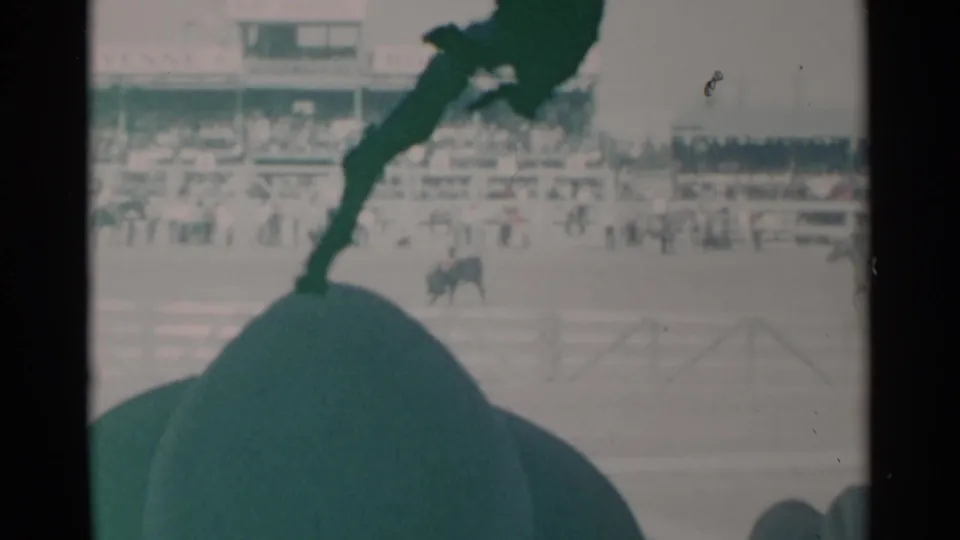 From a crowd seat in 1960 Cheyenne, the camera records a distant bull-riding moment with arena fencing and spectators behind.