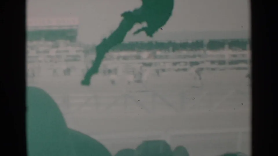 The home-movie frame shows rodeo action in Cheyenne with a horse and rider partly obscured by dark foreground shapes, grandstands, and arena rails.