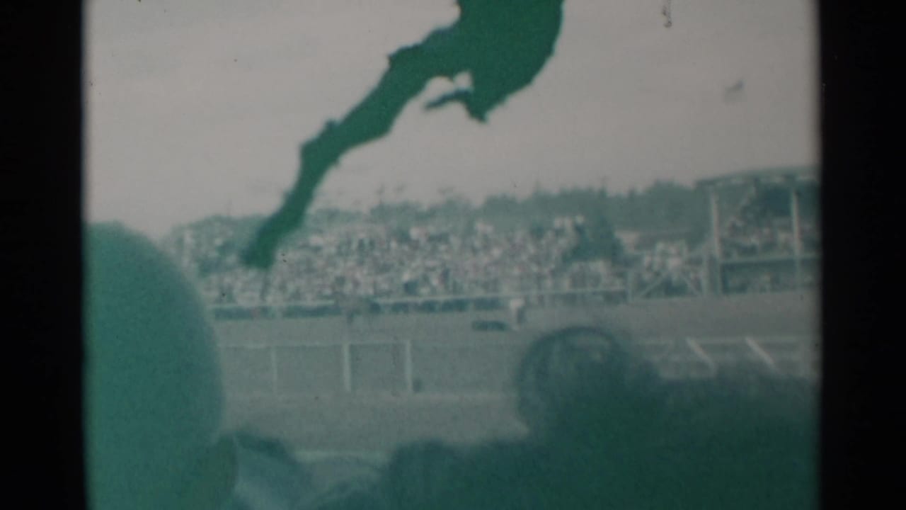 1960 Cheyenne home-movie frame showing two horses racing past distant bleachers, partly obscured by foreground spectators.