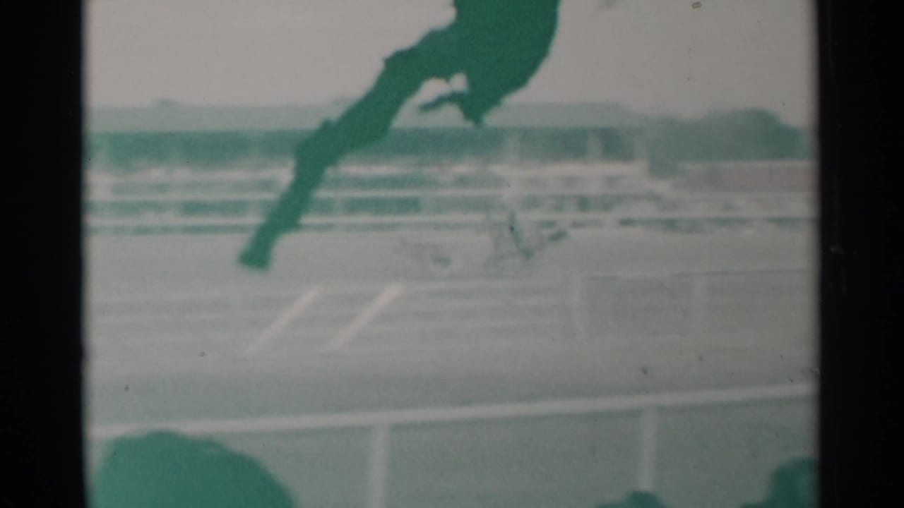 Racing horses pass along the track with railings and a grandstand in the background.