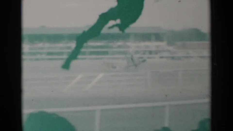 1960 Cheyenne 8mm frame of horses running on a dirt racetrack, with railings, grandstand seating, and soft spectator shapes.