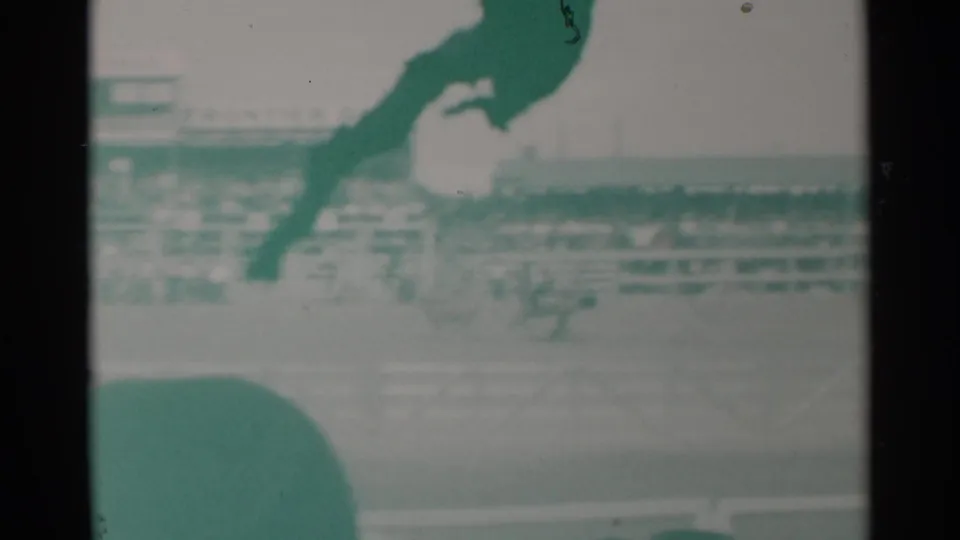 Soft green-tinted 1960 Cheyenne frame showing two horses on the dirt track, blurred crowd seating, and a spectator silhouette below.