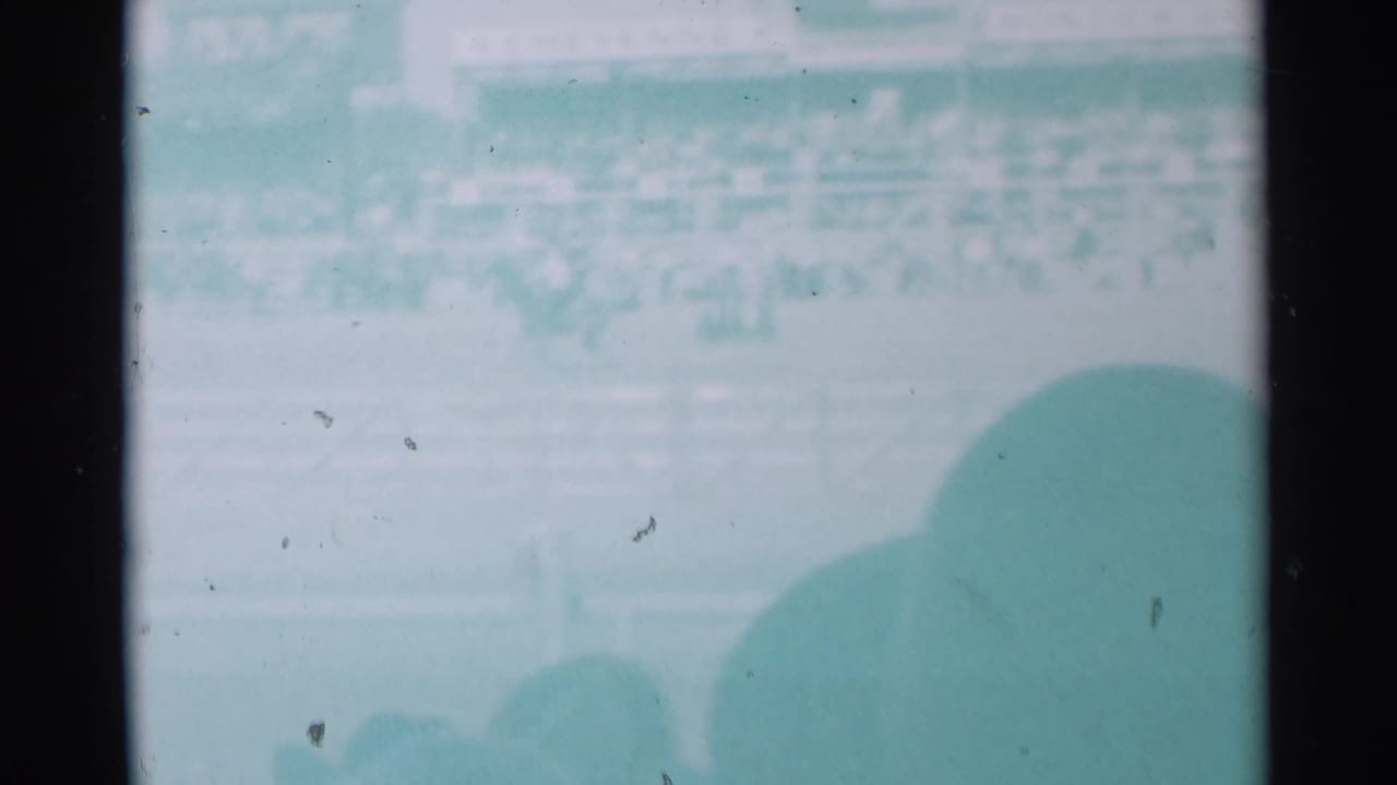 Faded view of a rodeo arena with railings, distant crowd, and spectator silhouettes in the foreground.