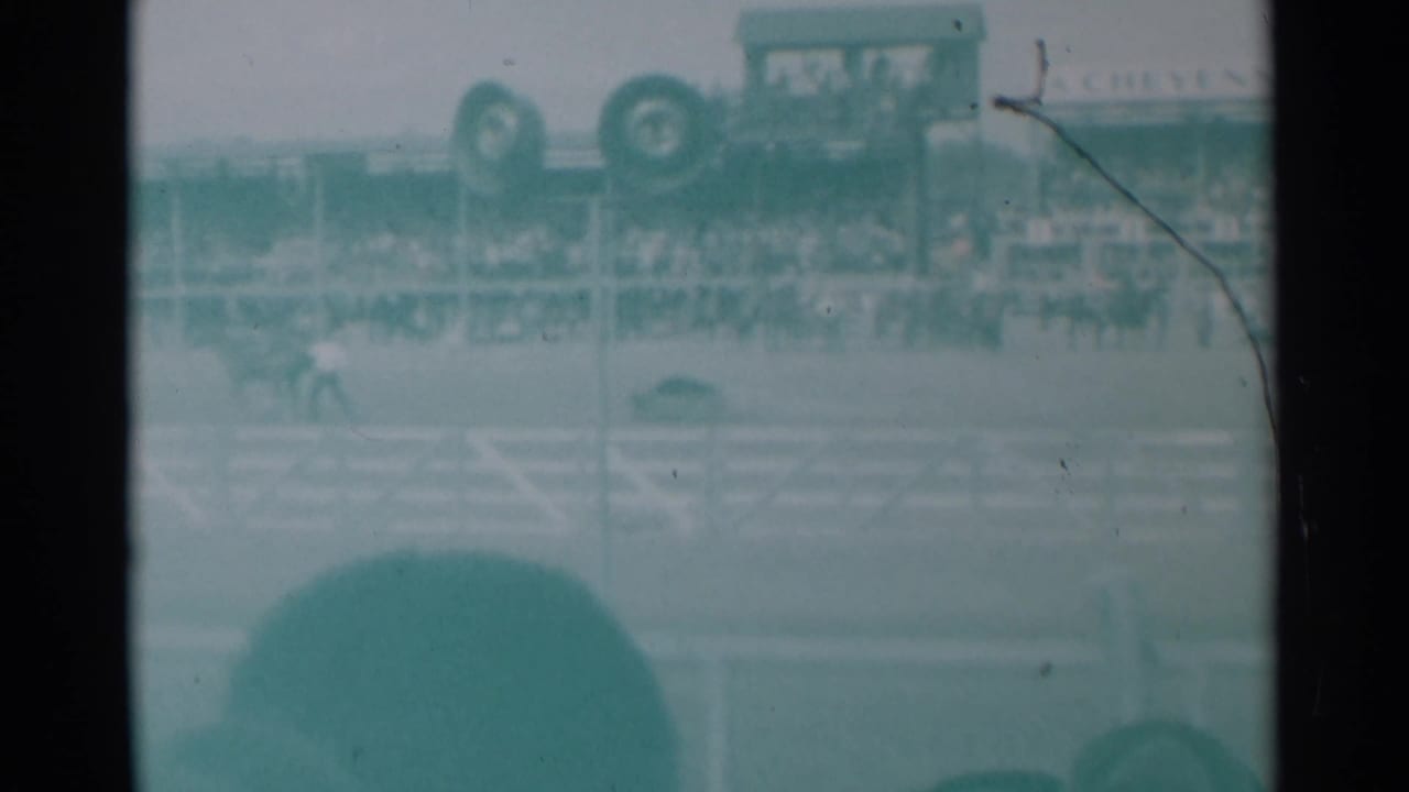 Rodeo arena with a rider near the rail, grandstand spectators, and vehicles or signage above the stands.