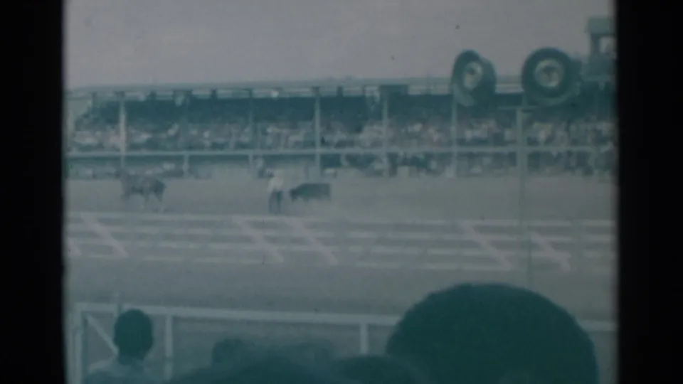 1961 Cheyenne rodeo footage frame showing horses in the arena, white fencing, crowded stands, and foreground spectator silhouettes.