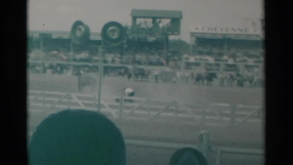 Cheyenne rodeo action continues in the arena, partly obscured by a foreground spectator's head and framed by grandstands and rail fencing.