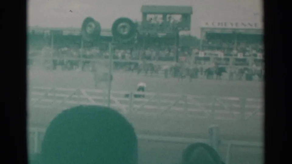 Soft 8mm frame of the Cheyenne rodeo arena, with a foreground spectator, white fencing, distant rider, crowd, and signage.