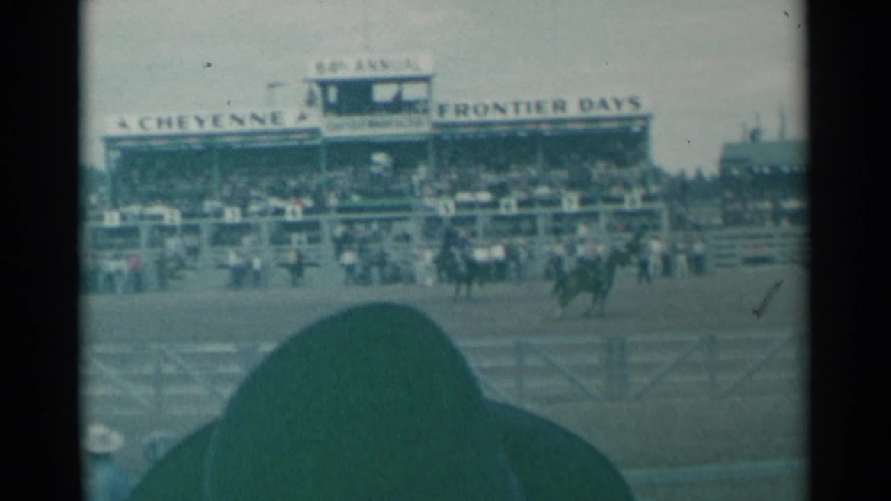 Rodeo arena with riders, grandstand signage, spectators, and a foreground hat.