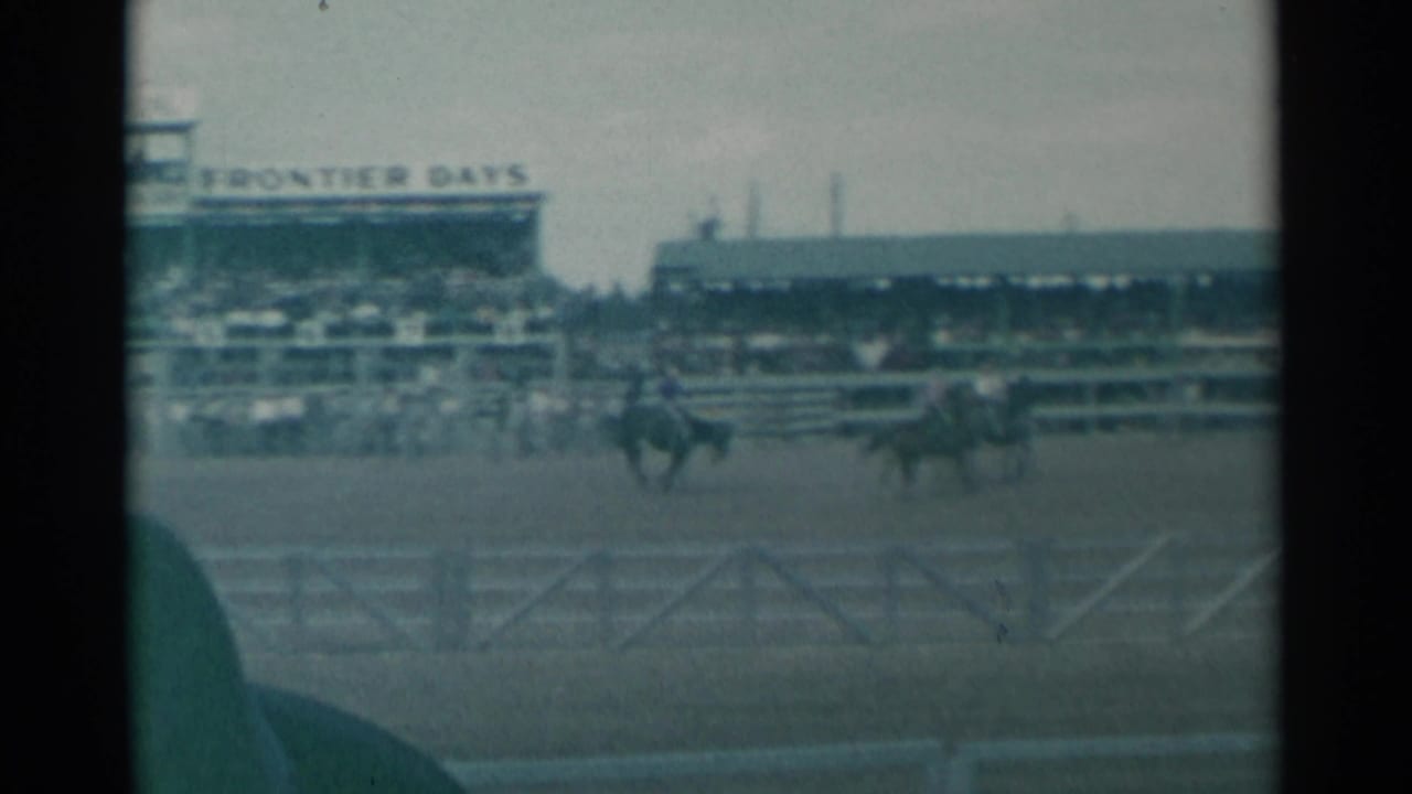 Two riders move across a rodeo arena in front of grandstands and fencing.