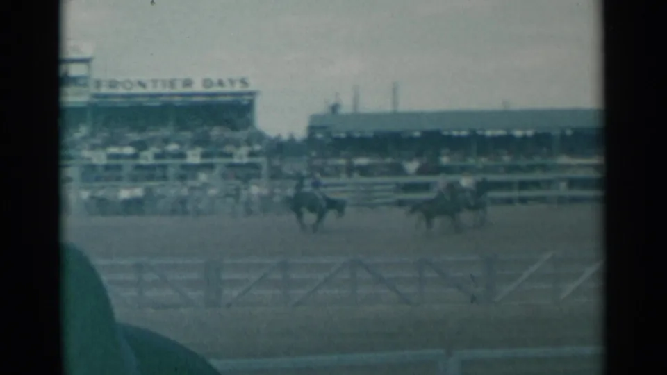 Riders on horseback pass through the Cheyenne Frontier Days arena, with spectators and long grandstand structures visible behind them.