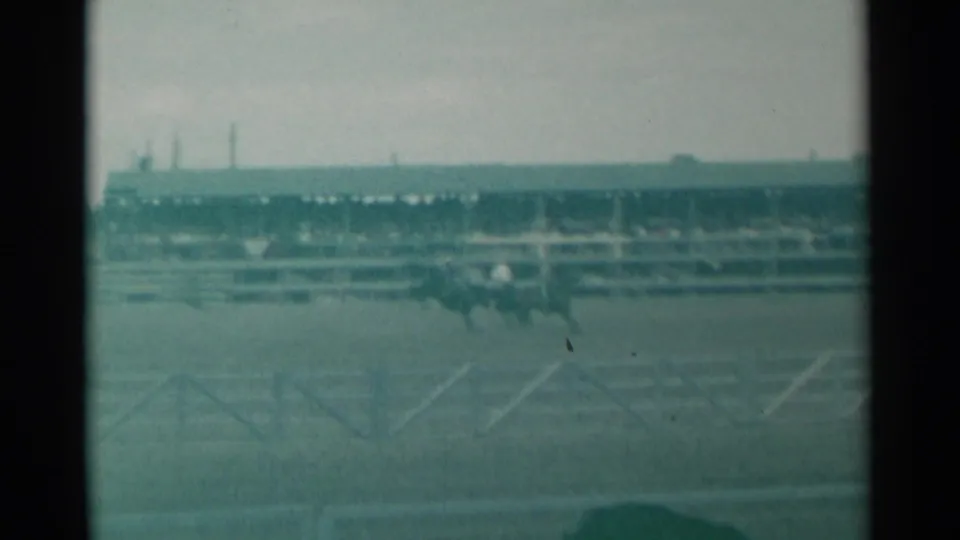 A soft, green-tinted 8mm view of the Cheyenne rodeo grounds shows distant riders, arena rails, and a long spectator grandstand.