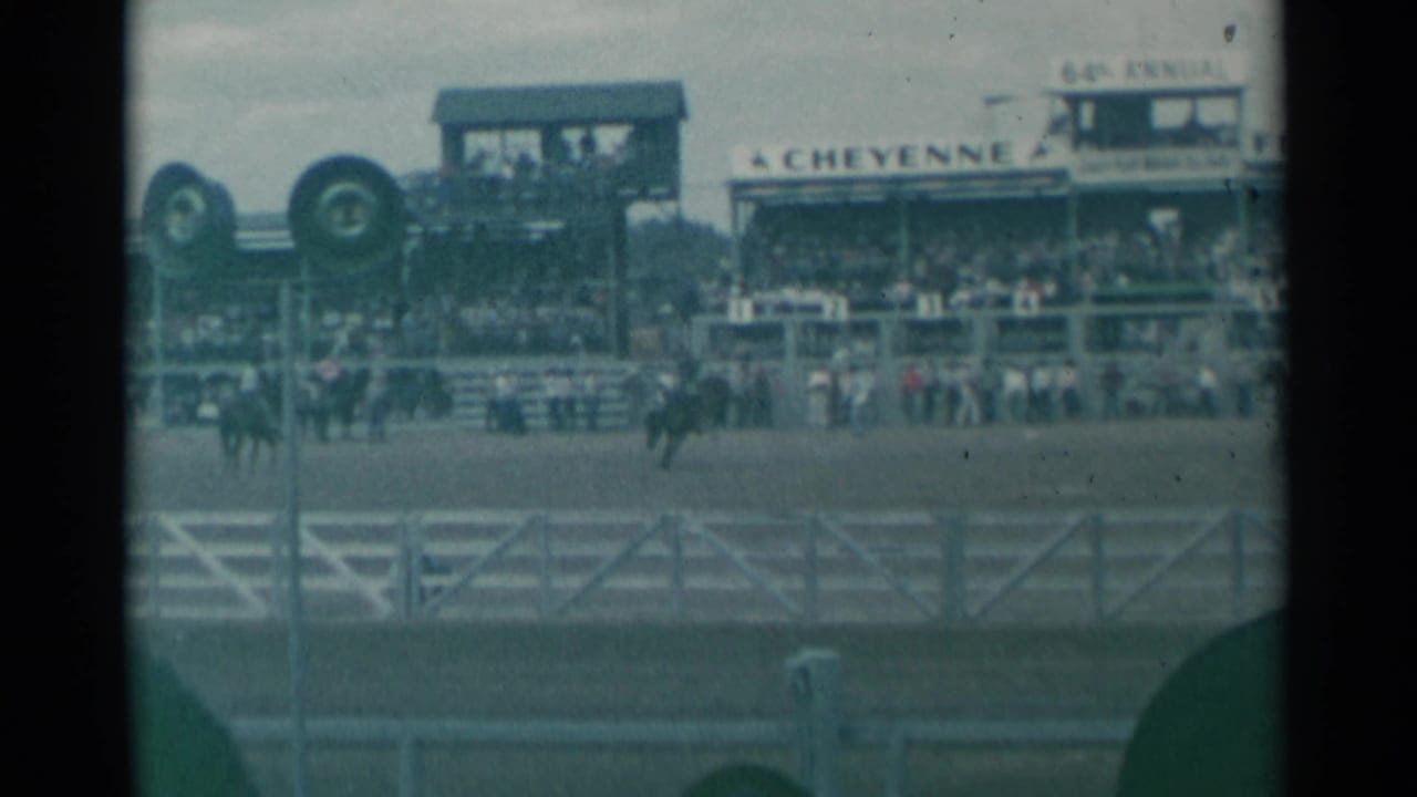 Riders on horseback move across a rodeo arena before crowded stands.