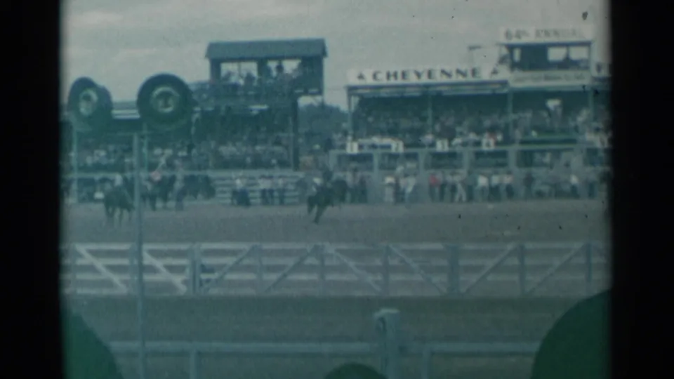 Spectator-shot 1961 Cheyenne rodeo frame showing mounted riders, arena fencing, and packed grandstands under a cloudy sky.