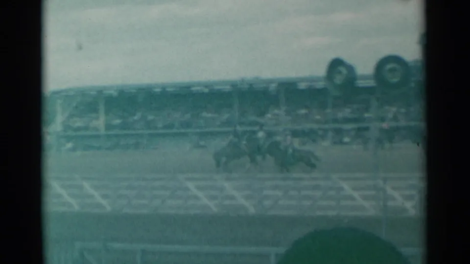 Wide 1961 Cheyenne rodeo frame with riders in the arena, white fencing, distant spectators, and event structures visible through soft film grain.