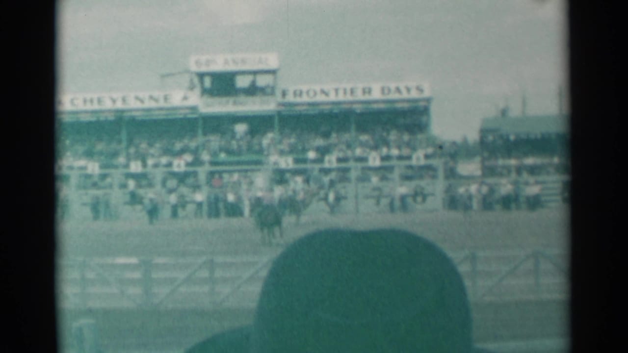 Rodeo arena with grandstand, spectators, signage, and a foreground hat partly blocking the view.