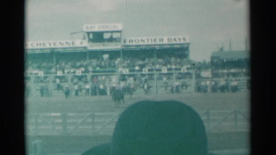 1961 Cheyenne, Wyoming rodeo arena seen from the stands, with Frontier Days signage, mounted riders, crowd, and a spectator hat in front.