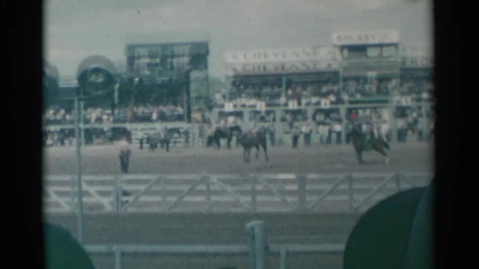 Visitor-shot 1961 Cheyenne rodeo frame showing riders in the arena, a packed grandstand, light poles, fencing, and foreground hats.