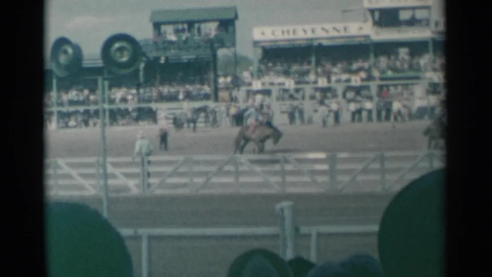 Bucking horse and rider at Cheyenne Frontier Days in 1961, filmed from behind spectators with grandstand signage visible.