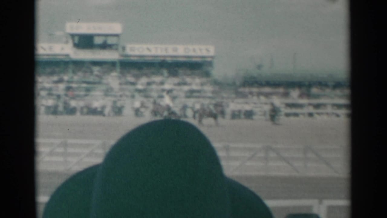 Rodeo arena with riders in the distance, grandstands, signage, and a dark foreground silhouette.