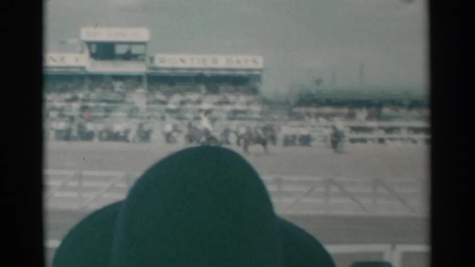 1961 Cheyenne rodeo arena view with distant horseback riders, crowded stands, visible signage, and a spectator silhouette in front.
