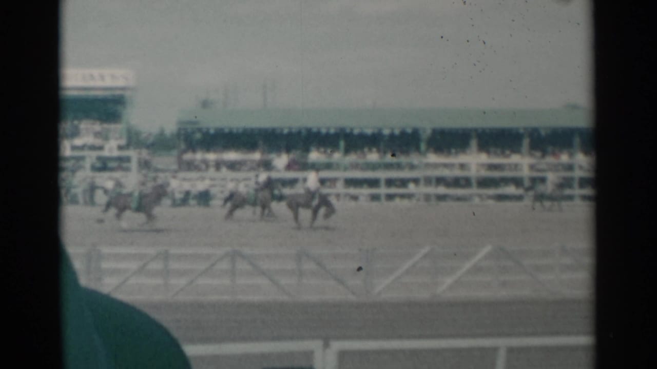 Distant riders on horseback move through the Cheyenne arena, with spectators seated behind the fence and a dark vertical film edge.