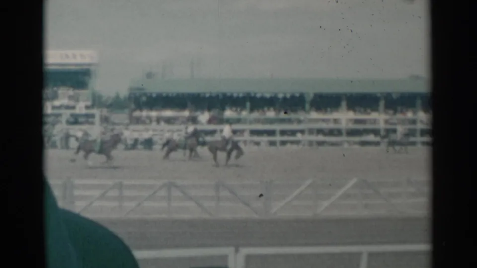 Distant riders on horseback move through the Cheyenne arena, with spectators seated behind the fence and a dark vertical film edge.