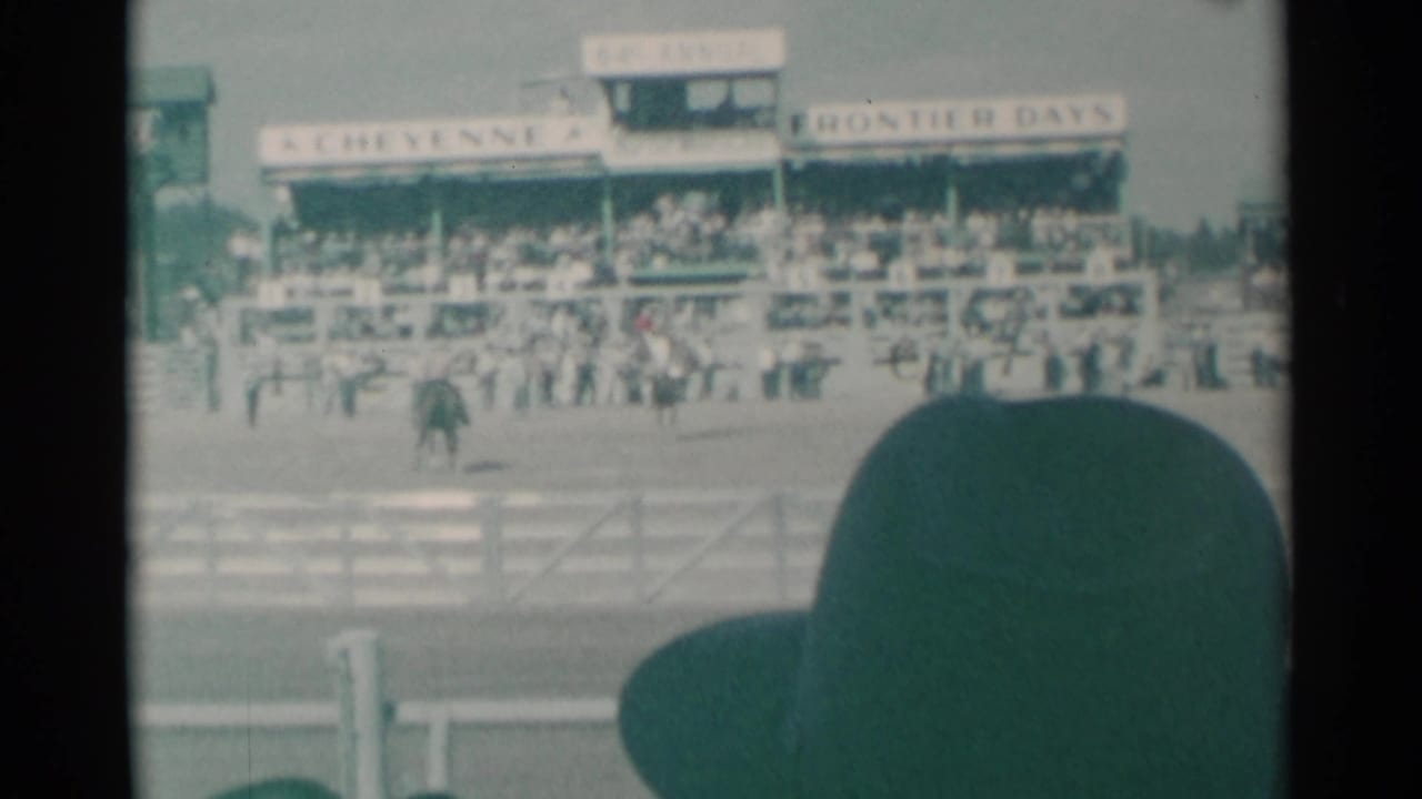 1961 Cheyenne rodeo scene viewed from the stands, with riders in the arena, crowded grandstands, white fencing, and foreground spectator hat.