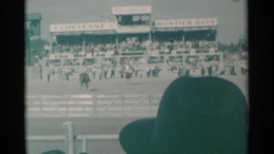 1961 Cheyenne rodeo scene viewed from the stands, with riders in the arena, crowded grandstands, white fencing, and foreground spectator hat.