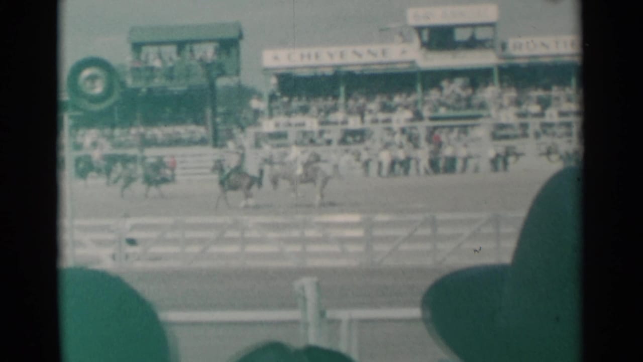 Mounted riders pass in front of grandstands and signage, partly blocked by foreground spectator silhouettes.