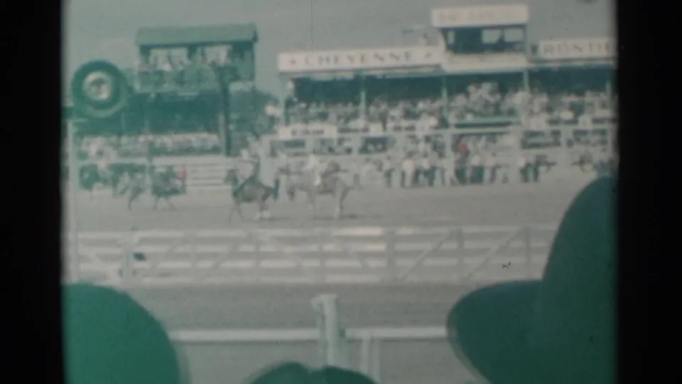 Cheyenne Frontier Days arena view with horseback riders moving across the frame, crowd-filled stands, event signs, and nearby spectators in silhouette.