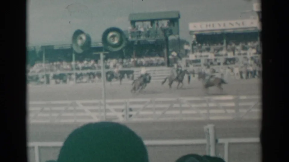 Distant 8mm view of a 1961 Cheyenne rodeo arena with riders and horses in motion, packed stands, arena fencing, and spectator hat blocking part of the frame.
