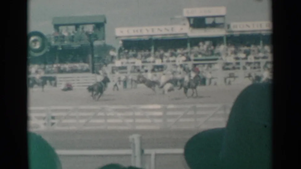 1961 Cheyenne Frontier Days rodeo footage showing multiple riders on horseback, a large audience, arena fencing, and foreground spectator silhouettes.