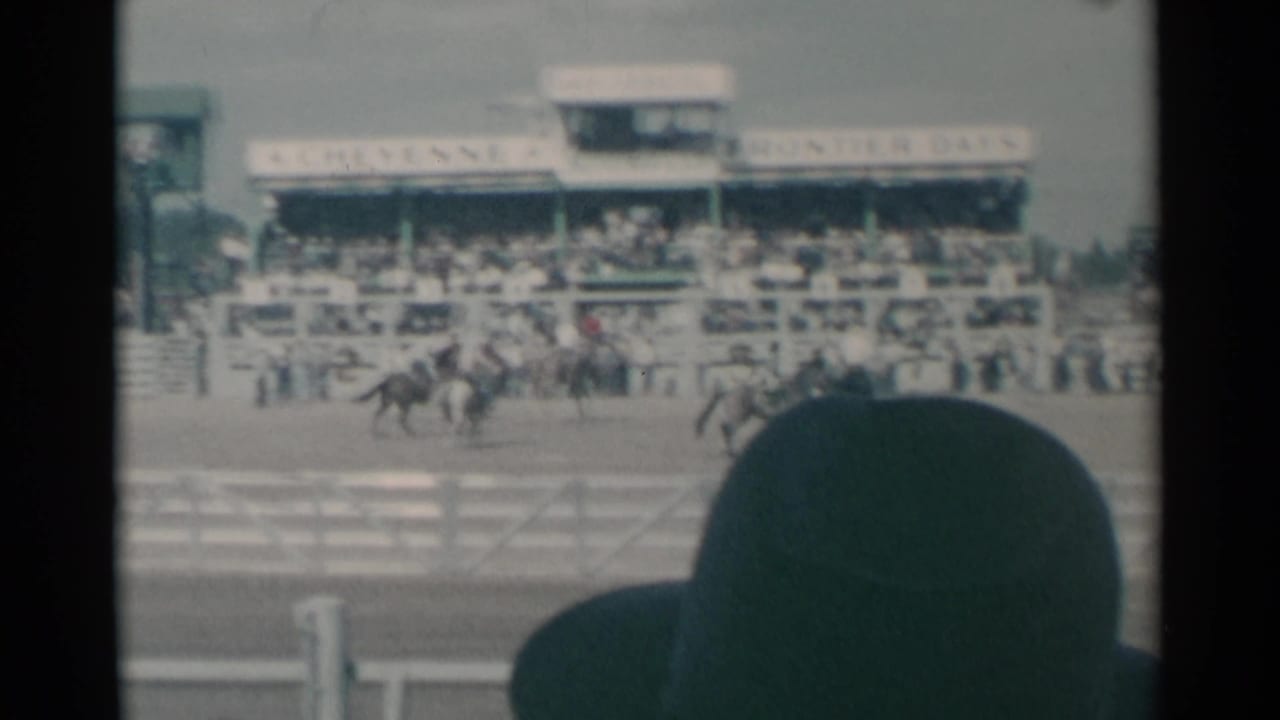 1961 Cheyenne rodeo view from the audience, with riders in the arena, crowded grandstand, and a foreground spectator partially blocking the frame.