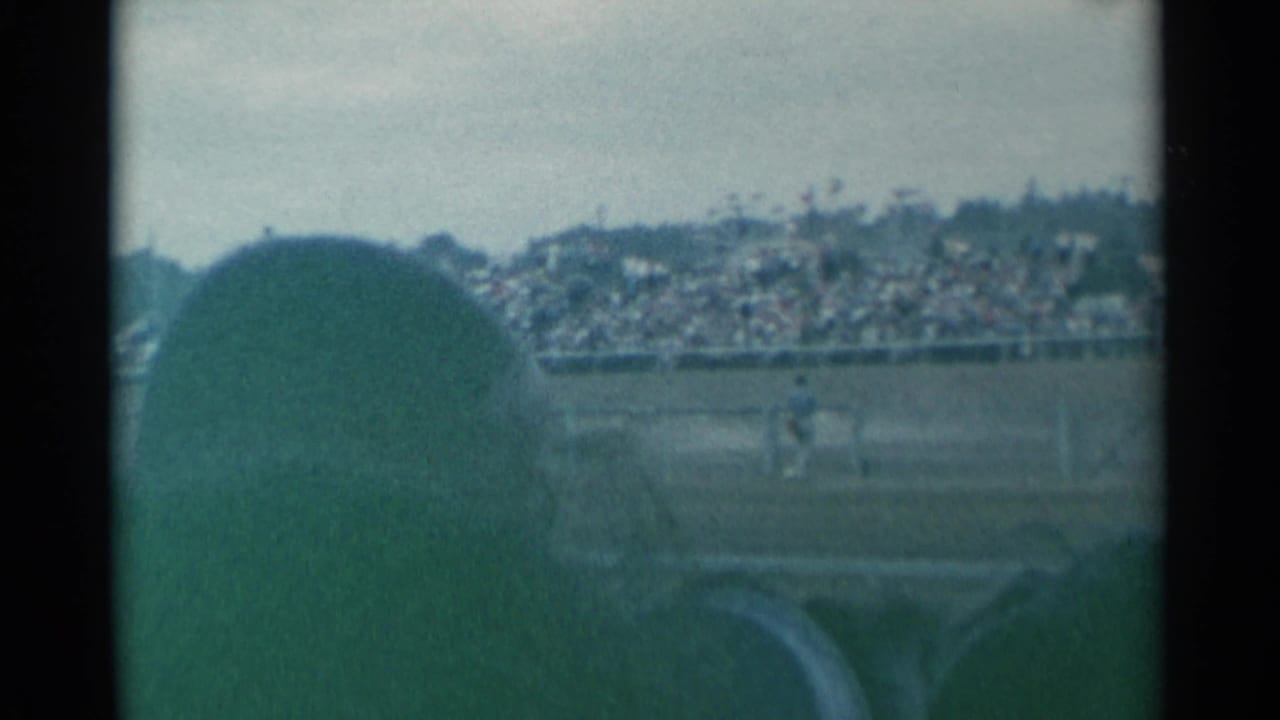 Foreground spectator silhouette faces a rodeo arena with distant grandstand and rider.