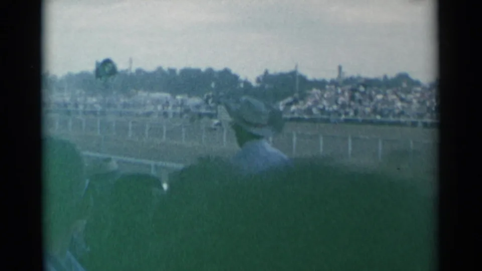 Cheyenne Frontier Days arena view with a foreground spectator hat, distant fencing, and crowded stands visible through faded 8mm color.