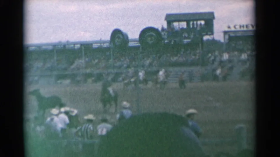 Home-movie frame from the Cheyenne rodeo showing riders in the arena, spectators in foreground seats, grandstand structures, and partial signage.