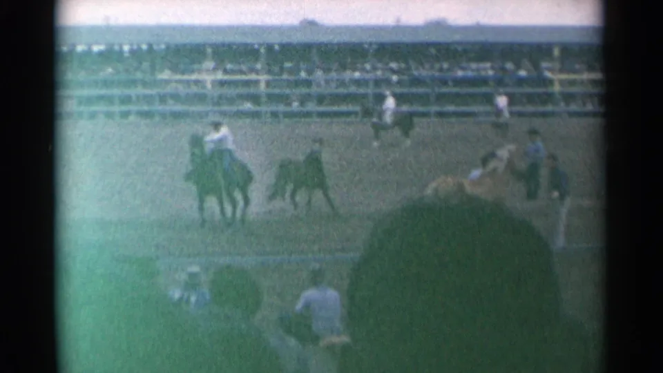 At the 1961 Cheyenne rodeo, a rider on horseback and nearby livestock are seen in the arena with workers and grandstands behind.