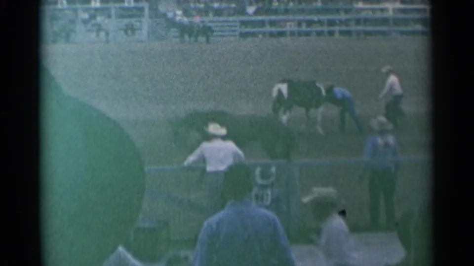 1961 Cheyenne rodeo scene with a horse in the arena, cowboys nearby, spectators behind fencing, and a foreground spectator partly blocking the frame.