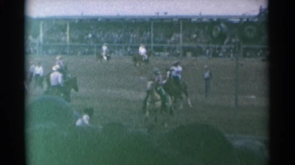 A rider appears on or near a bucking horse in the Cheyenne rodeo arena while cowboys and spectators watch from the field and stands.