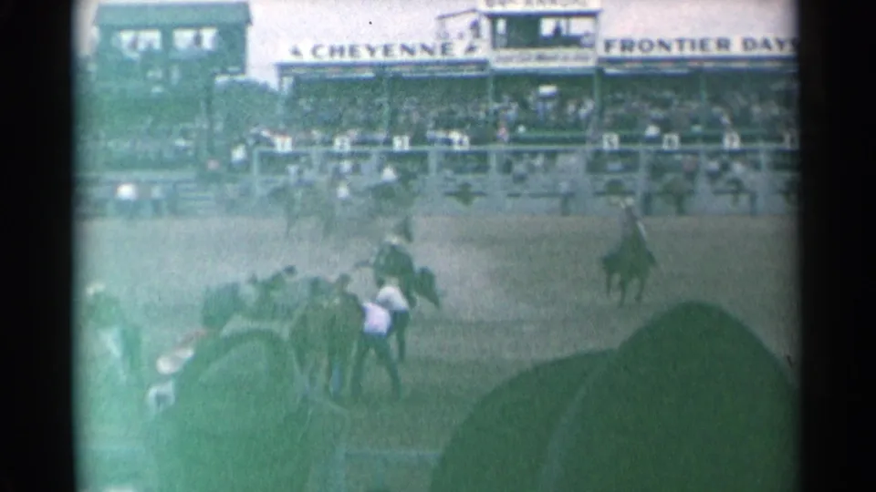 Rodeo arena beneath a visible Cheyenne Frontier Days sign, with cowboys, horses, rail fencing, and spectators in the grandstand.